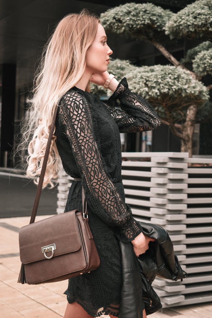 Stylish young woman posing outdoors in a black lace dress with a leather bag.
