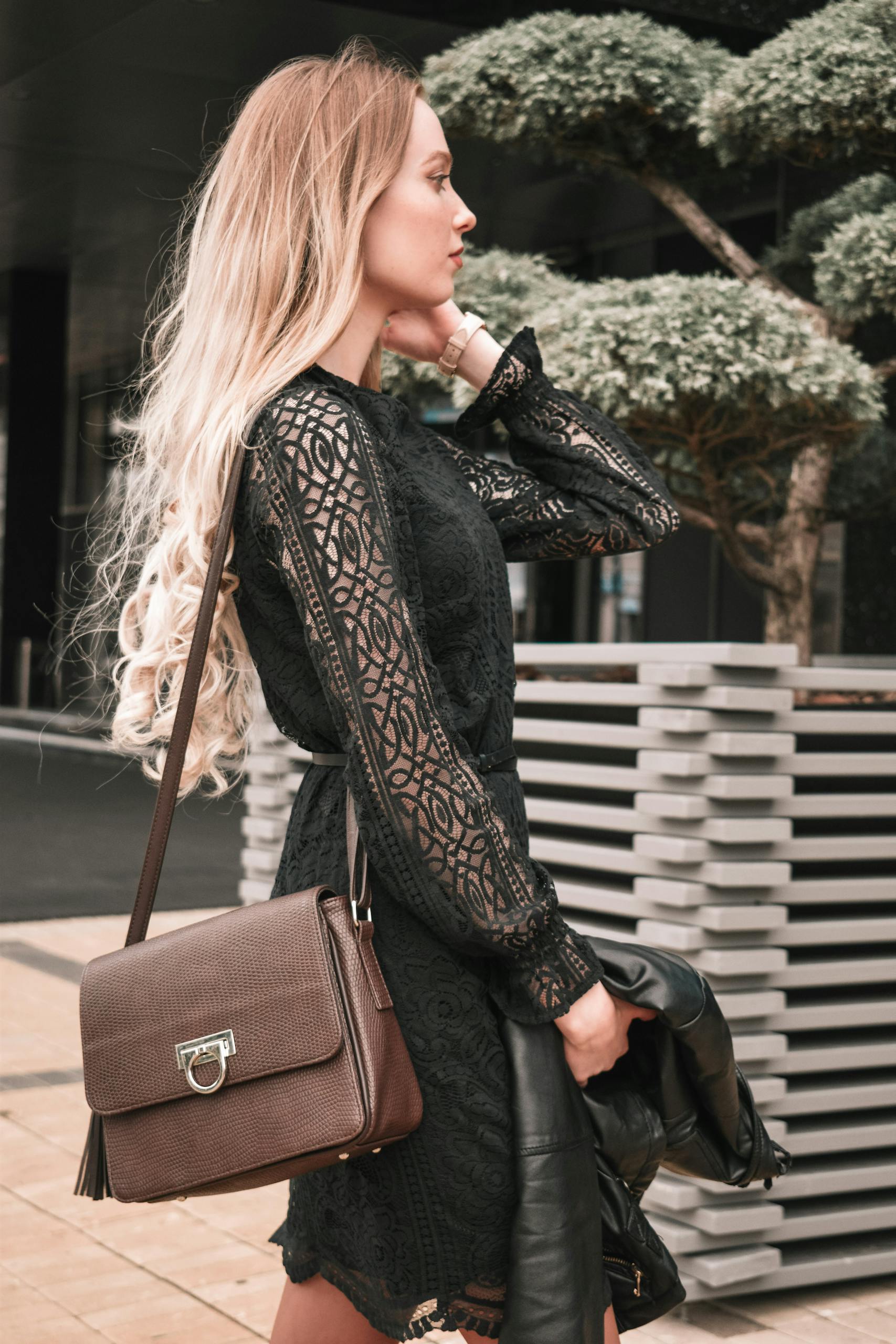 Stylish young woman posing outdoors in a black lace dress with a leather bag.